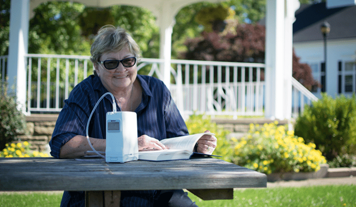 Happy and healthy elderly woman at the park reading a book with her OxyGo portable oxygen concentrator.